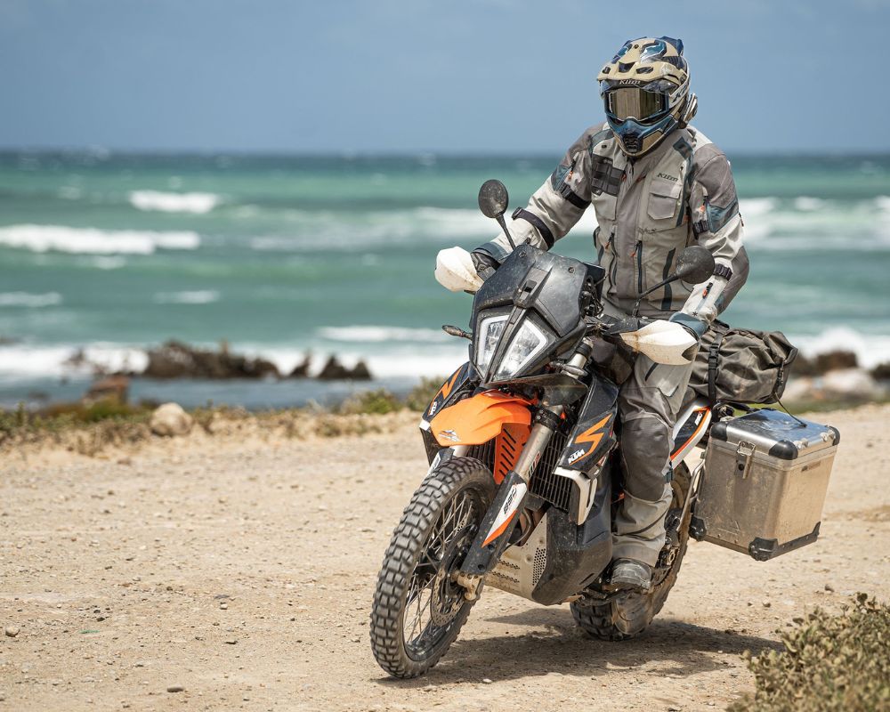 Rider wearing Klim Badlands Pro A3 jacket and pants on a KTM adventure motorcycle near the ocean