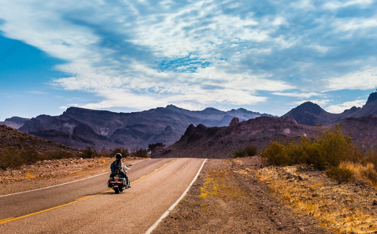 Motorcyclist riding through a desert highway surrounded by Arizona mountains under a partly cloudy sky