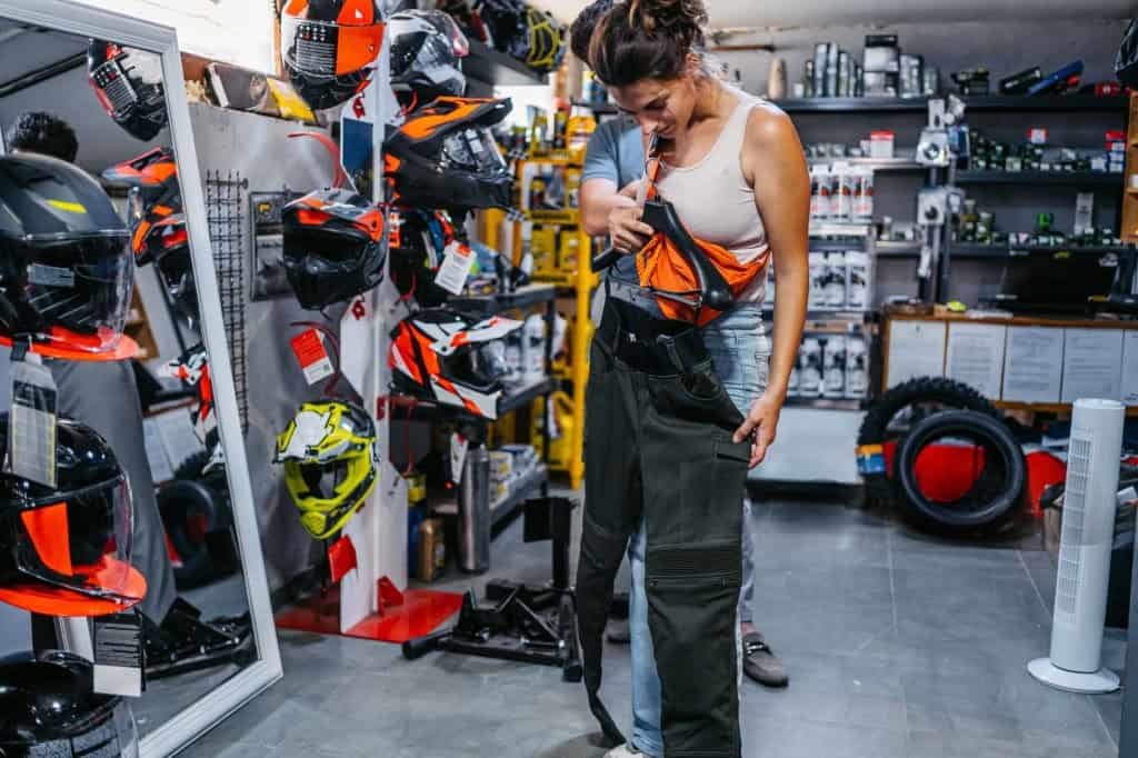 Woman trying on motorcycle pants in a gear shop surrounded by helmets and riding accessories.