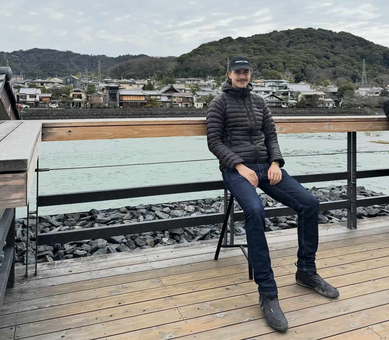 Man sitting by a riverside deck wearing casual motorcycle gear and protective riding shoes