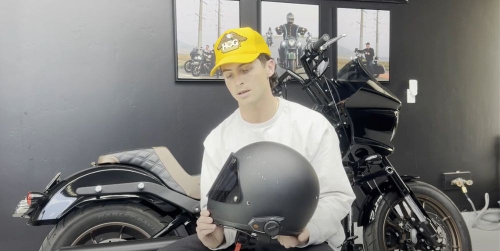 Rider holding a matte black Biltwell Gringo helmet with a Sena communicator installed, seated next to a black motorcycle.