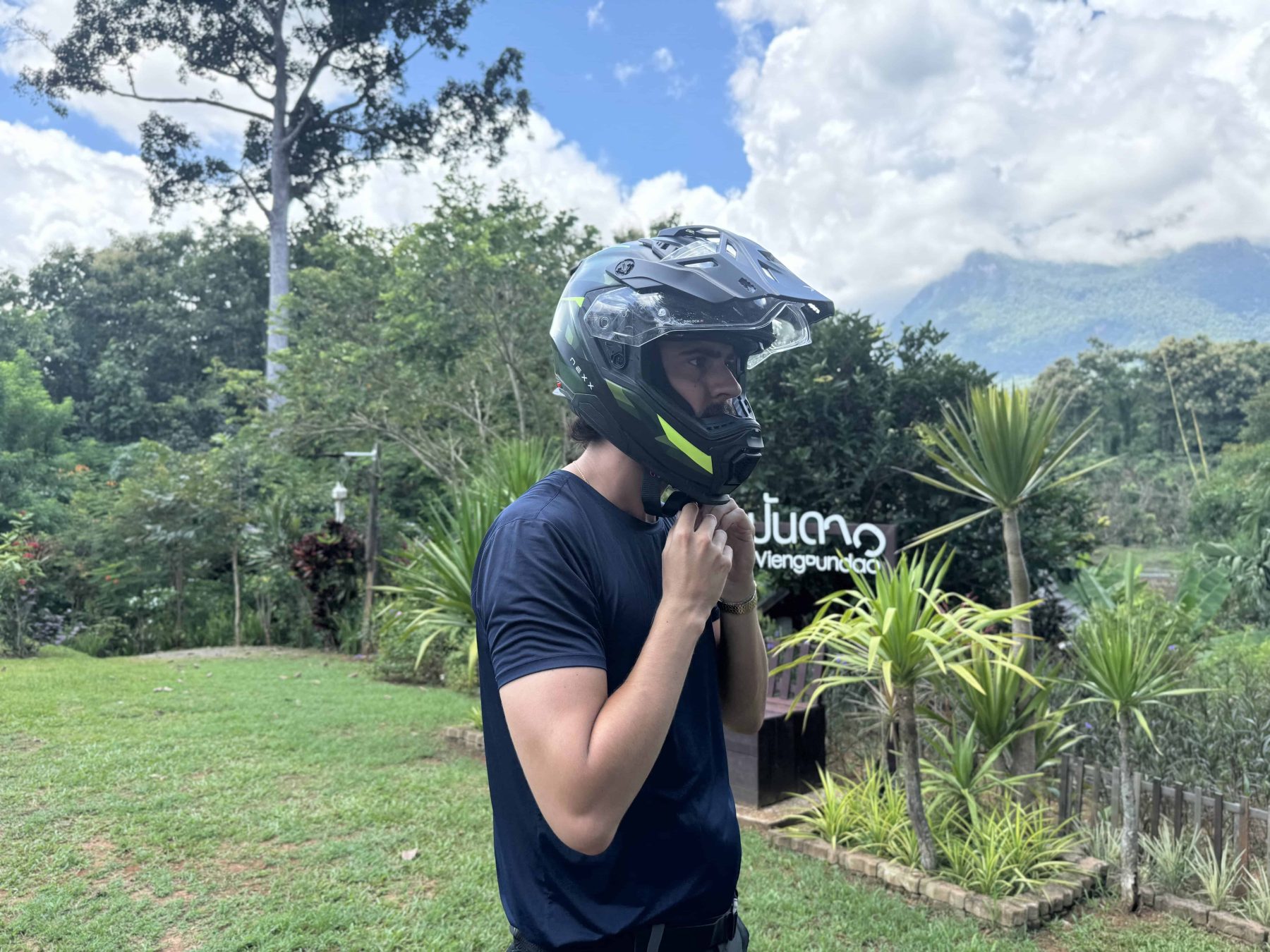 Close-up of a rider wearing the NEXX X.WED3 helmet, showcasing the chin strap and peak design in a tropical outdoor setting.