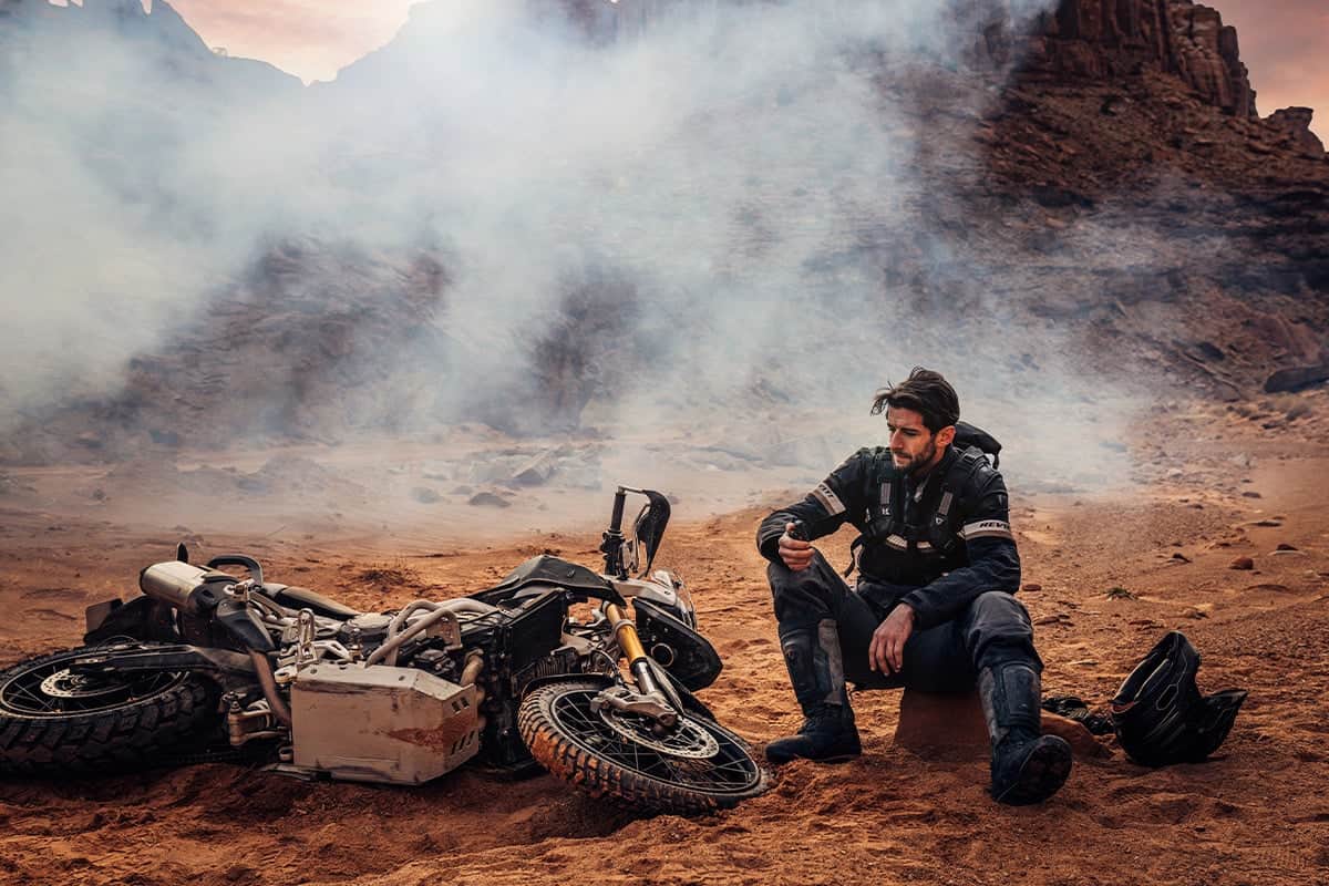 Motorcyclist sitting injured in remote desert terrain beside fallen bike, holding a GPS or communication device