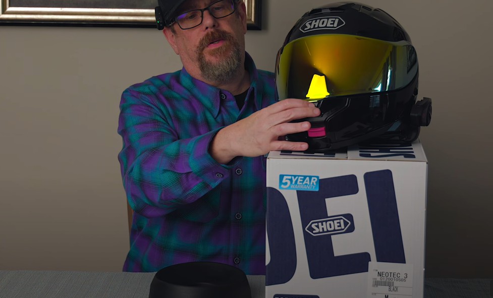 Hand demonstrating the new center locking mechanism on a black Shoei Neotec 3 helmet resting on its Shoei-branded box.