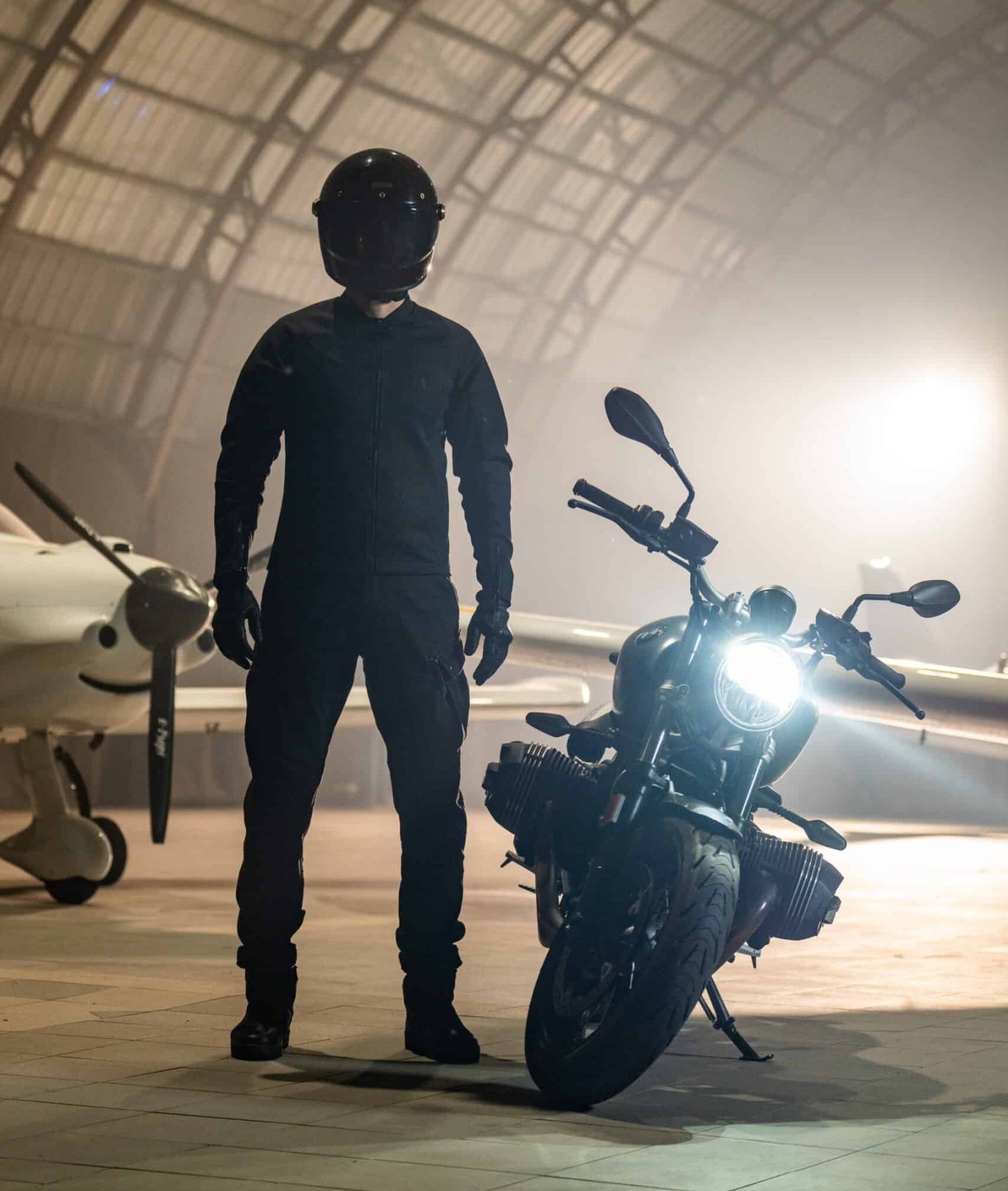 Rider in full black gear standing next to a motorcycle in a hangar, showcasing the Pando Moto Mark AAA jeans in a modern tapered fit