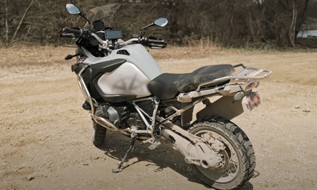 Adventure motorcycle with GPS mounted on handlebars, parked on a dirt trail in a forested area