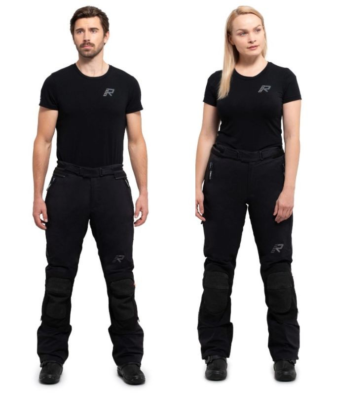Male and female riders wearing Rukka Armatou-R waterproof motorcycle pants, shown standing front-facing in full gear.