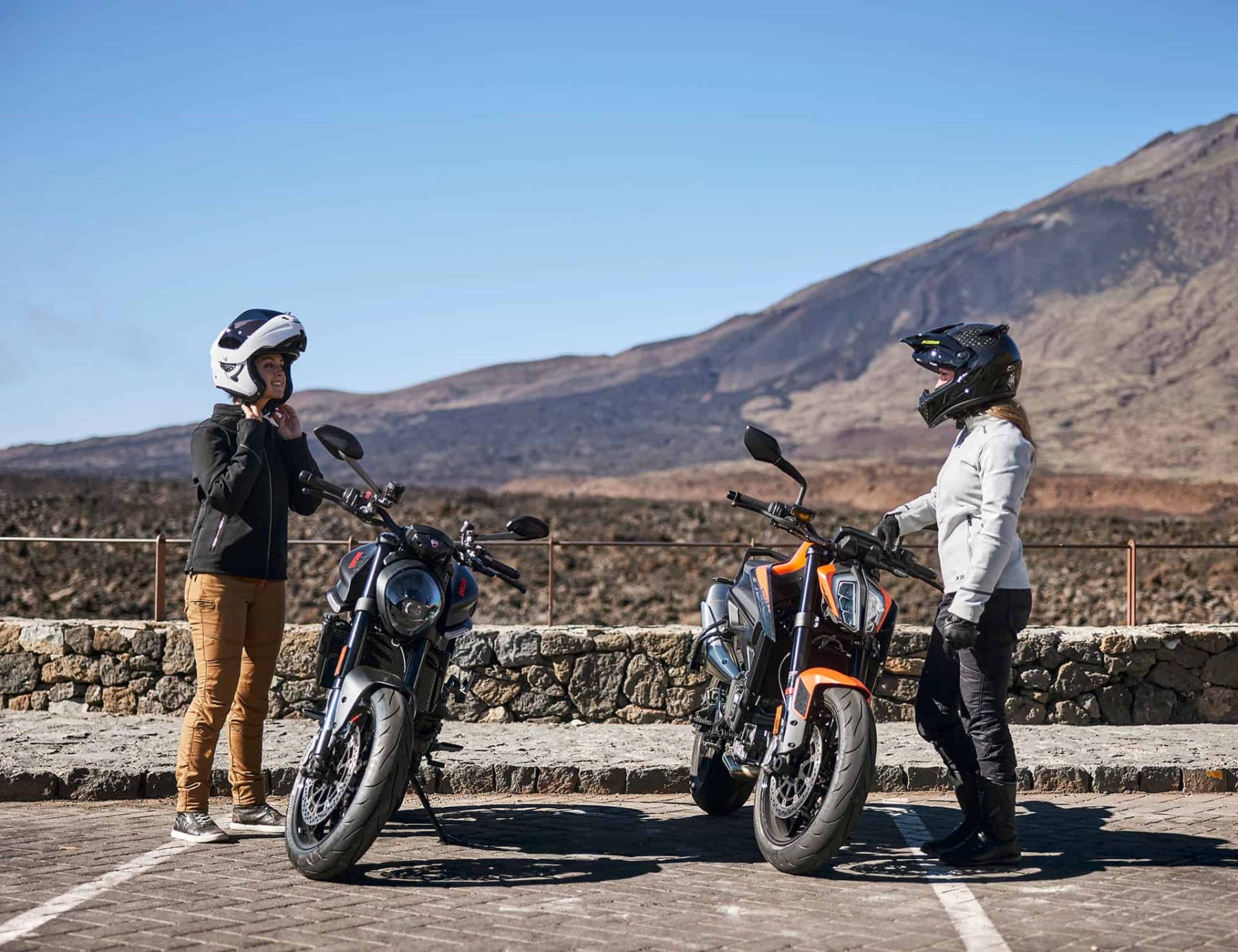 Two women riders standing beside their motorcycles in protective gear, wearing female-specific riding pants and jackets