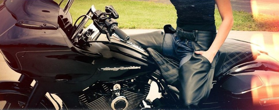 Woman in leather chaps sitting on a black Harley-Davidson motorcycle, with a casual cruiser-style outfit.
