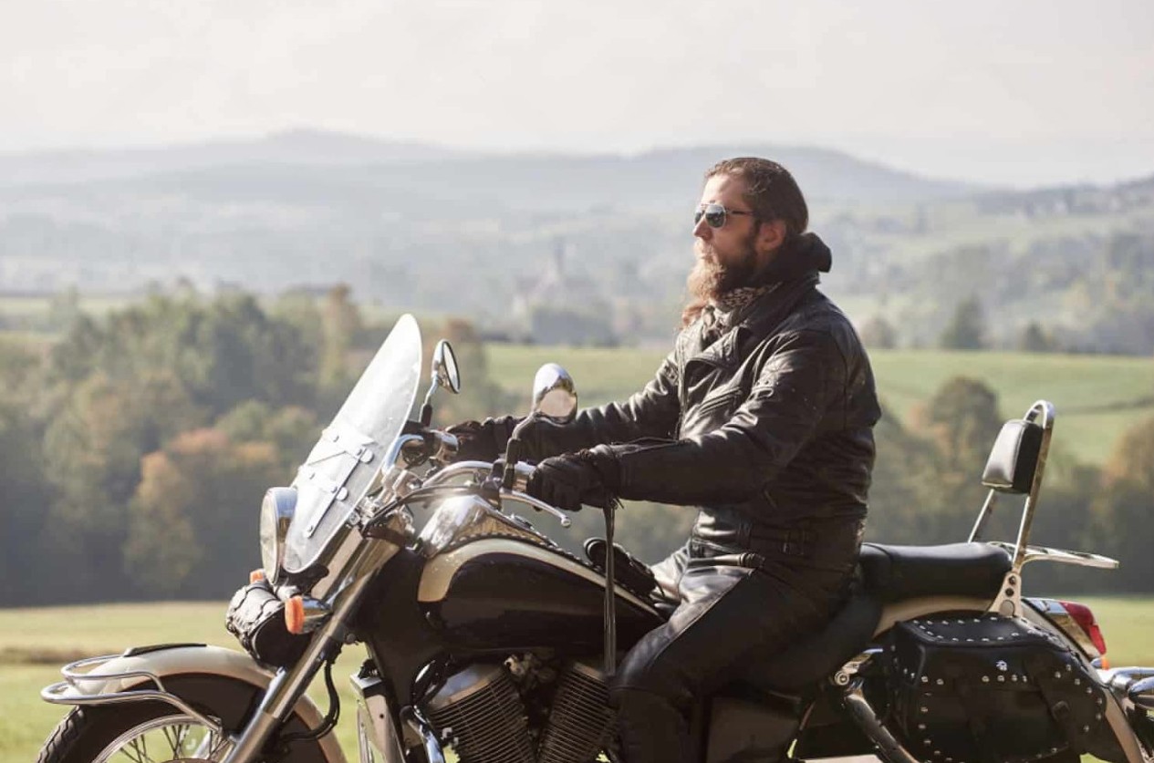 Motorcyclist wearing a full leather outfit, including pants and jacket, riding through a scenic countryside.