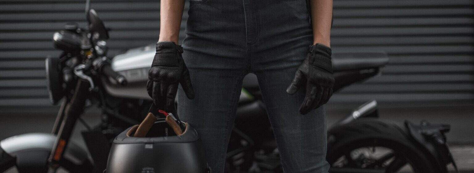 Man in a black leather motorcycle jacket and gloves standing confidently near a modern metal structure, embodying a vintage-meets-modern style.