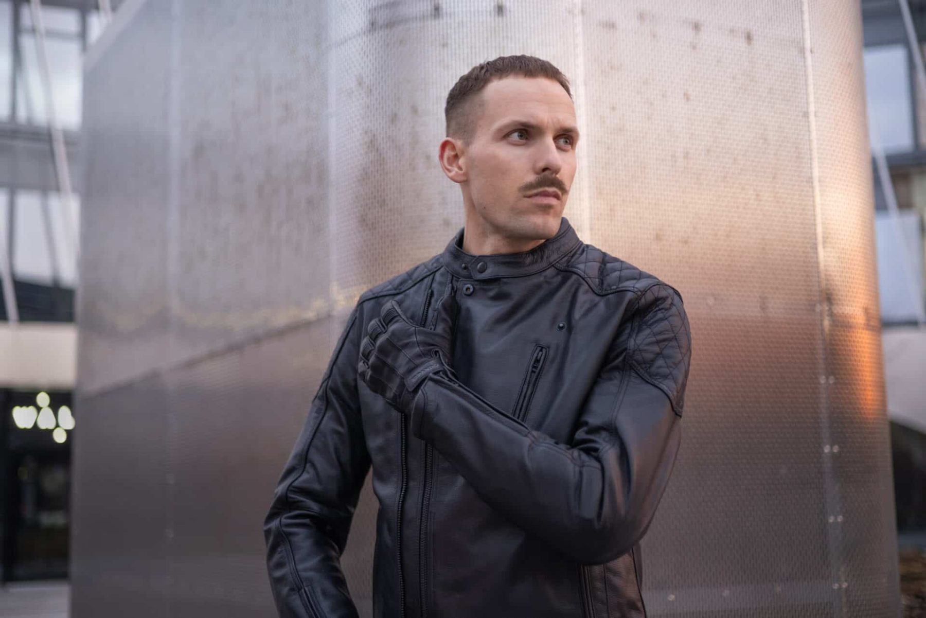Man in a sleek black motorcycle jacket and gloves standing in front of a metal wall, showing modern gear with classic styling.