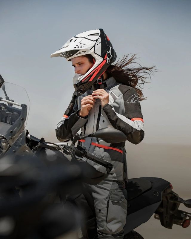 Female rider zipping up her Alpinestars Stella Andes V3 jacket while standing next to an adventure motorcycle in a desert environment.