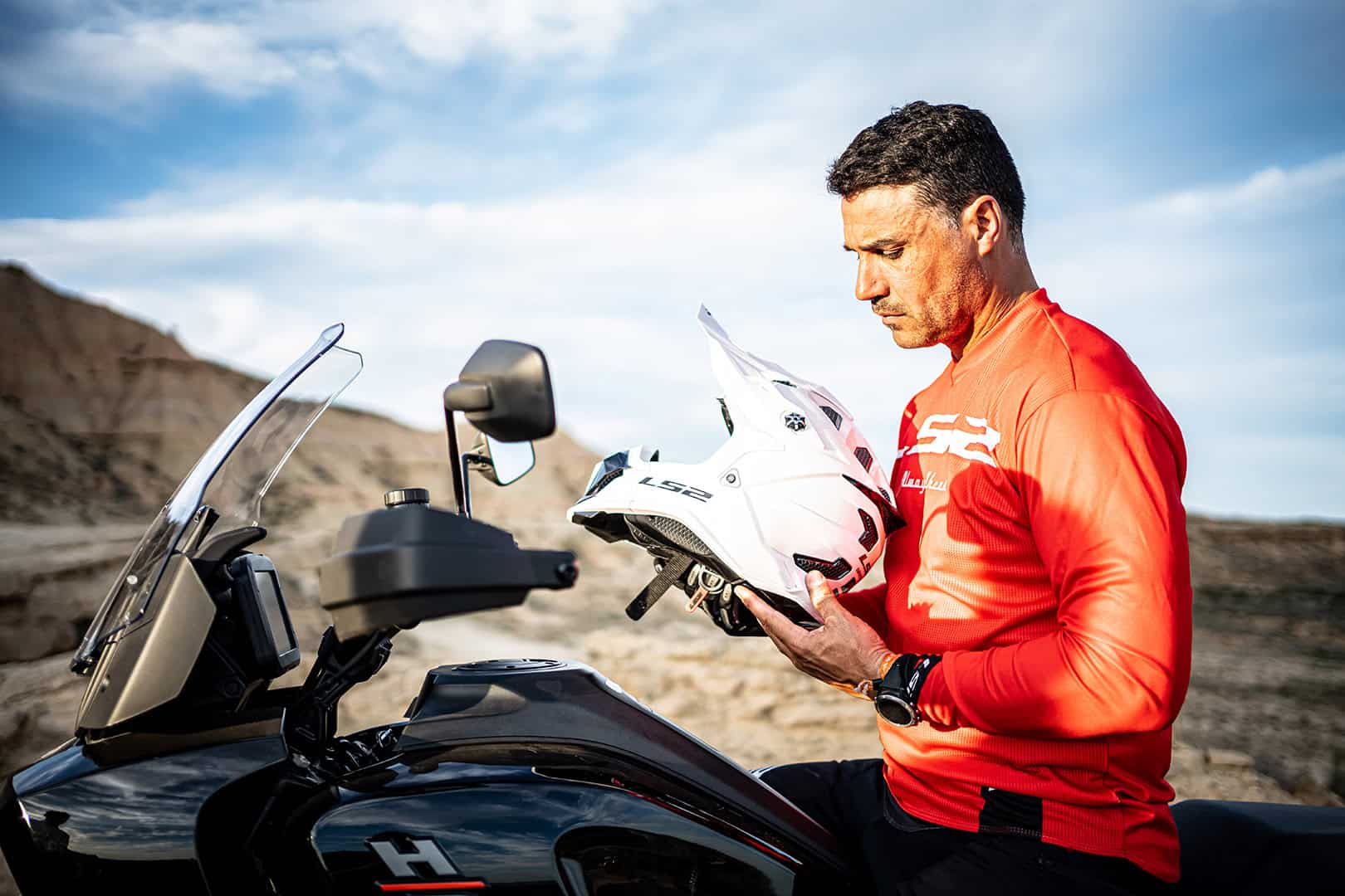 Rider in red jersey holding LS2 off-road helmet beside touring motorcycle in desert landscape