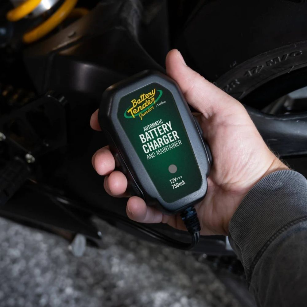 Battery Tender Junior 12V, 750mA charger held in hand beside a motorcycle.