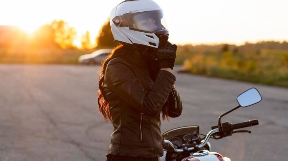 Female motorcyclist at sunset wearing a white helmet and black leather jacket, preparing to ride