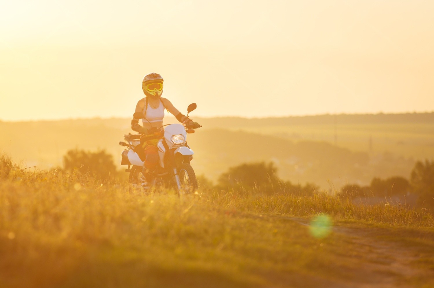 Motorcyclist riding through a sunny open field at golden hour, dressed in warm-weather riding gear on a white dual-sport motorcycle.