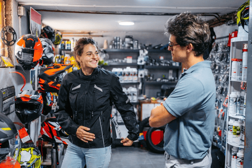 Woman trying on a motorcycle jacket at a gear shop, discussing fit and options with a store associate surrounded by helmets and accessories.
