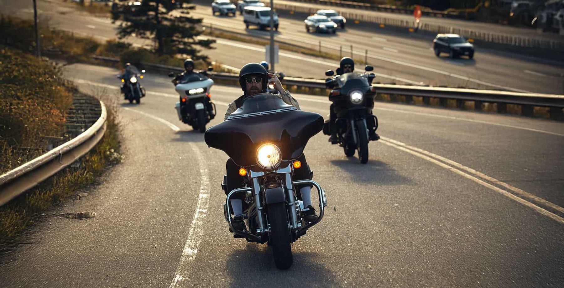 Group of motorcyclists riding cruisers on a highway exit ramp, led by a rider wearing a half helmet and using communication gear