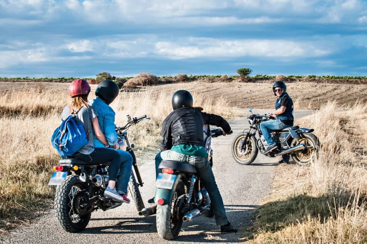 Group of motorcyclists stopped on a rural road in summer, wearing casual riding gear and helmets, chatting during a warm-weather ride.