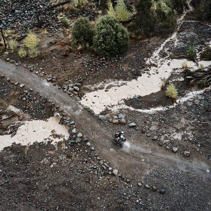 Aerial view of two adventure motorcyclists pausing on a remote rocky trail surrounded by rugged terrain and streams.