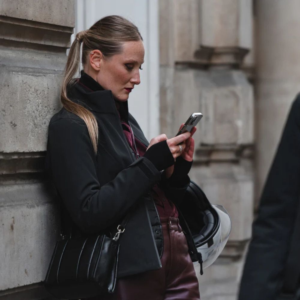 Woman in a fitted motorcycle jacket leaning against a wall, holding a phone and helmet—showcasing tailored fit and mobility off the bike.