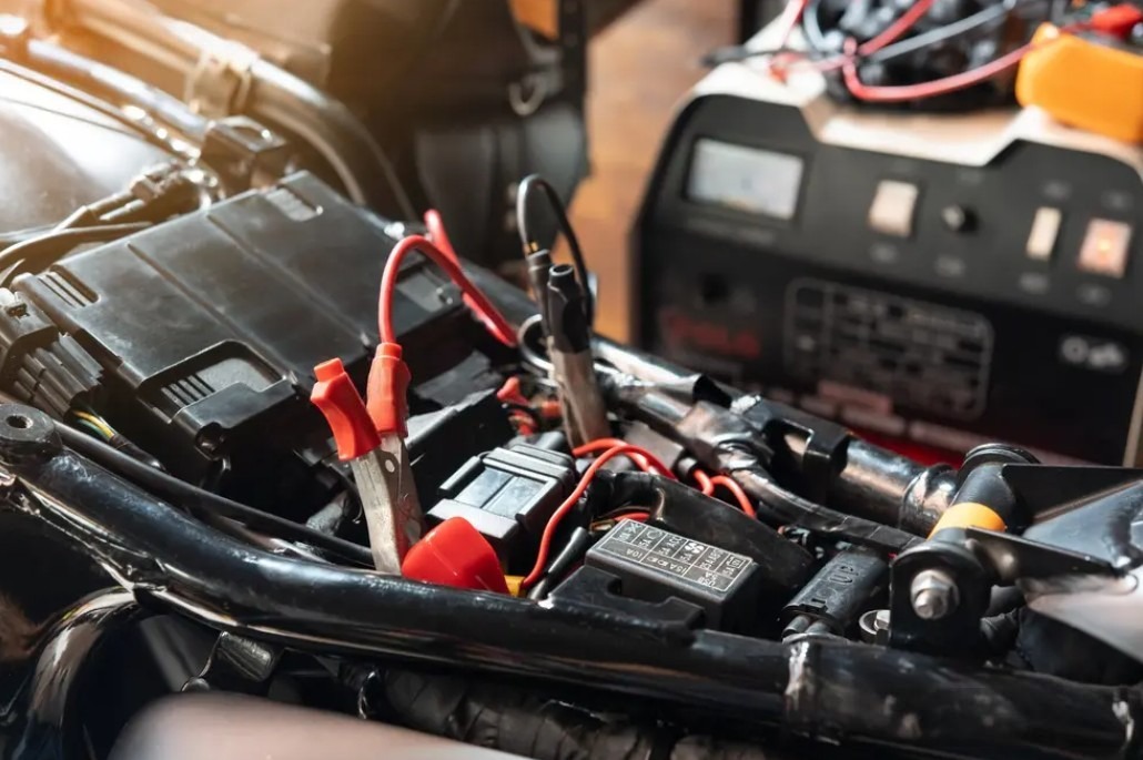 Motorcycle battery charging in progress with red and black clamps connected and a smart charger in the background.