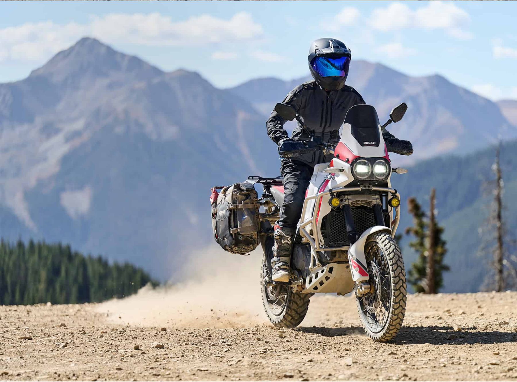 A woman riding a Ducati adventure bike in full Klim Sedona gear on a rocky trail with mountains in the background