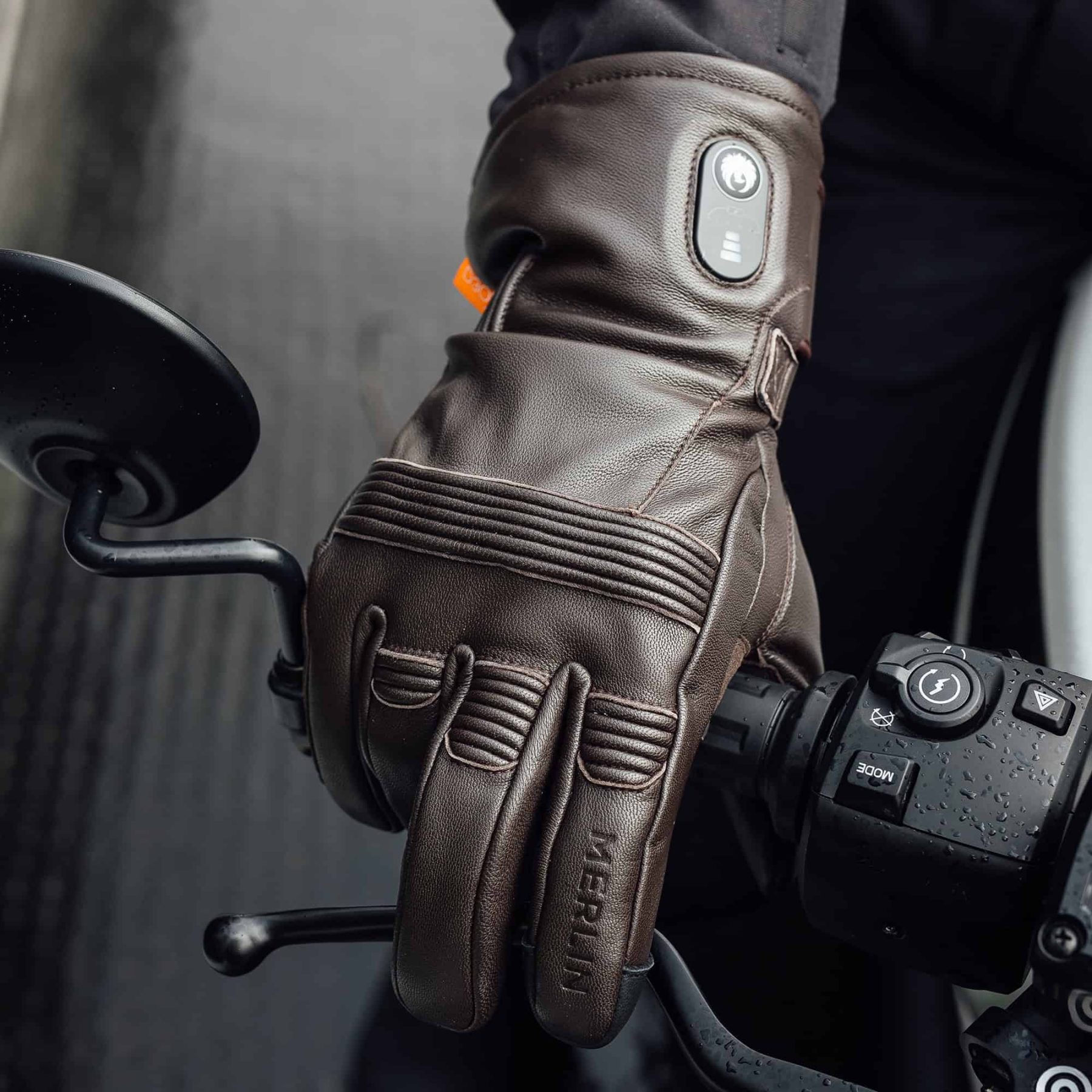Close-up of a rider wearing brown Merlin Minworth II heated gloves gripping a motorcycle handlebar on a rainy day.