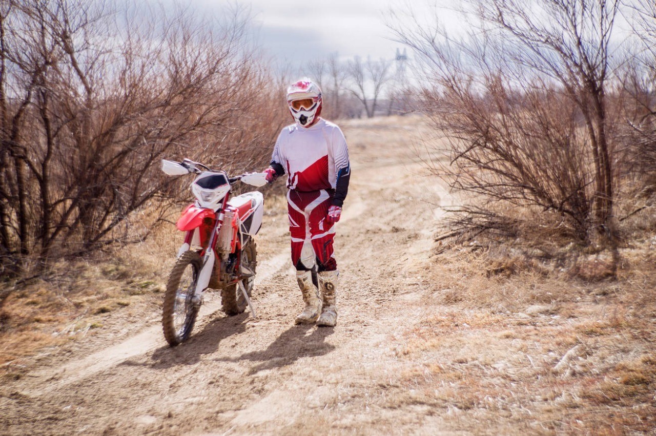 Rider in motocross gear walking a dirt bike through a dry off-road trail, wearing stiff, protective MX boots.