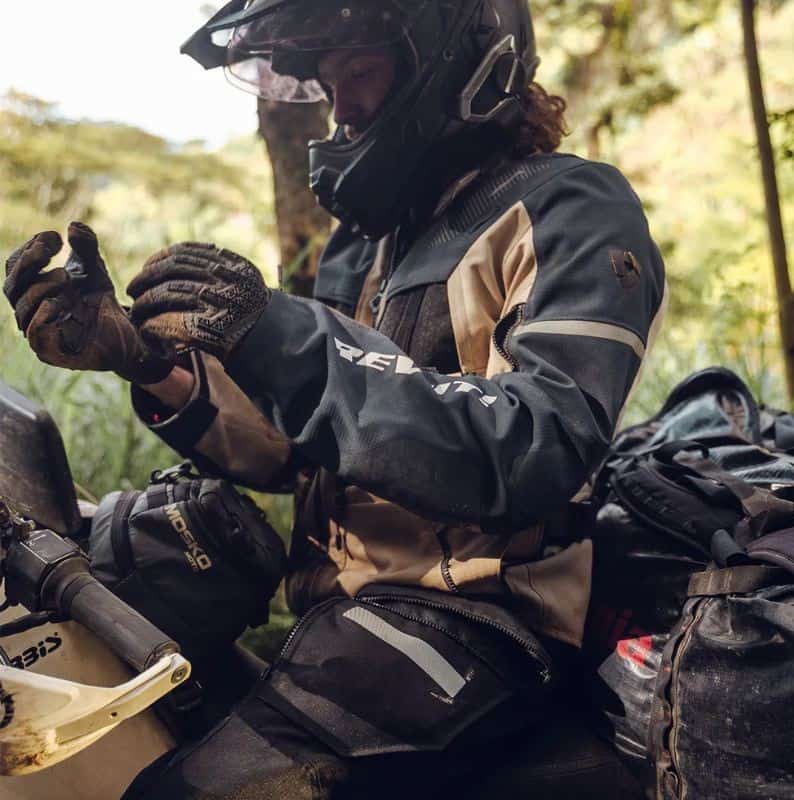 Adventure rider wearing REV’IT! gear adjusting gloves while seated beside a dual-sport motorcycle in the forest