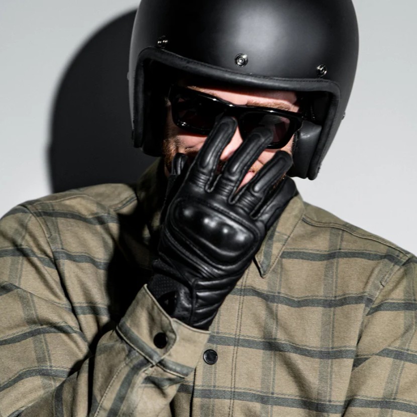 Rider wearing black leather motorcycle gloves and helmet, covering face with gloved hand