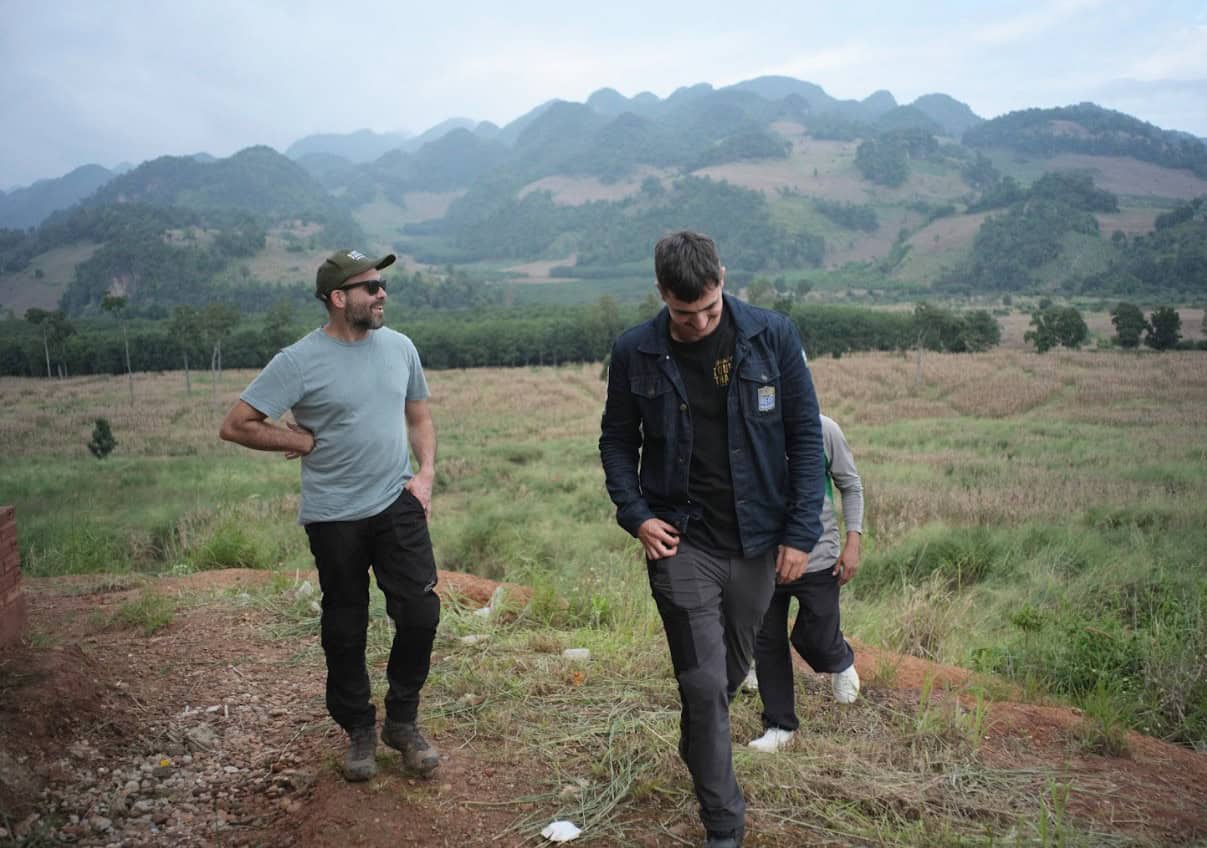 Three riders in casual motorcycle gear walking through a grassy, rural landscape with scenic mountains in the background