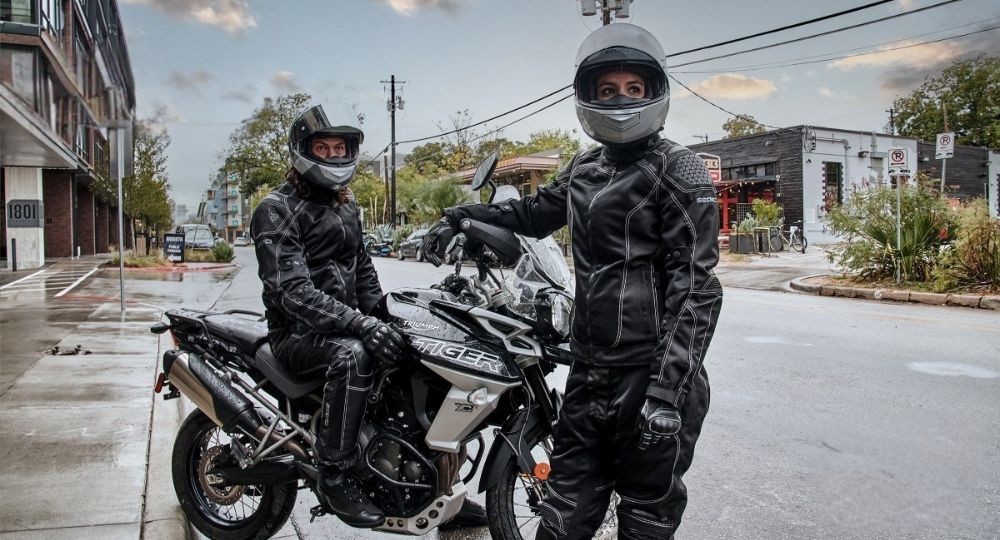 Two riders wearing full motorcycle gear, including the Sedici Alexi 2 Mesh Women's Jacket, standing with a Triumph Tiger on a wet city street.