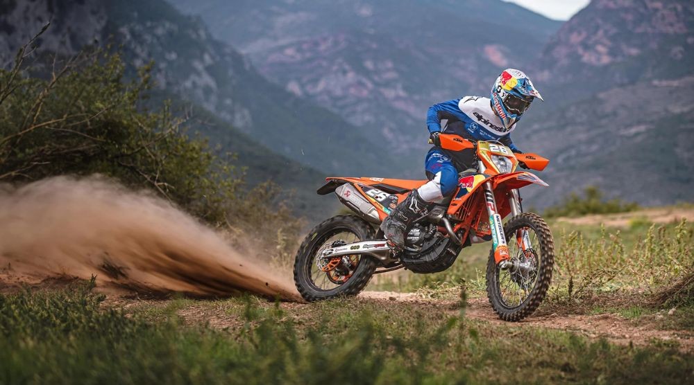 Rider wearing full protective motocross boots powers through a dusty off-road trail on an orange KTM bike in mountainous terrain