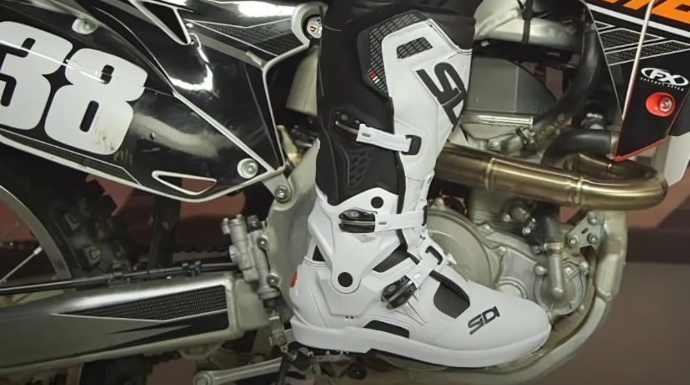 Close-up of a rider wearing white Sidi Atojo SRS boots mounted on a dirt bike, showing the boot’s slim profile and hinge system.