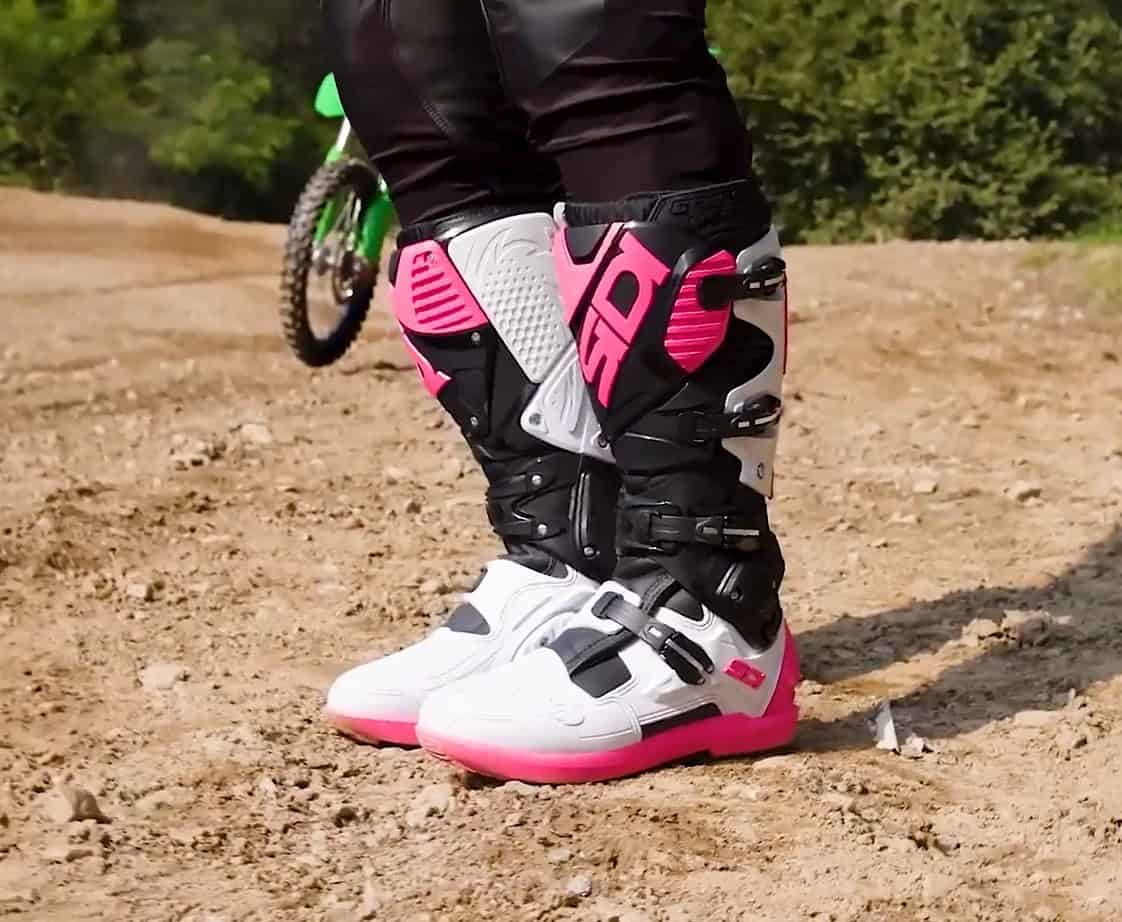Close-up of Sidi Crossfire 3 SRS motocross boots in white, black, and pink colorway, worn on a dirt trail.