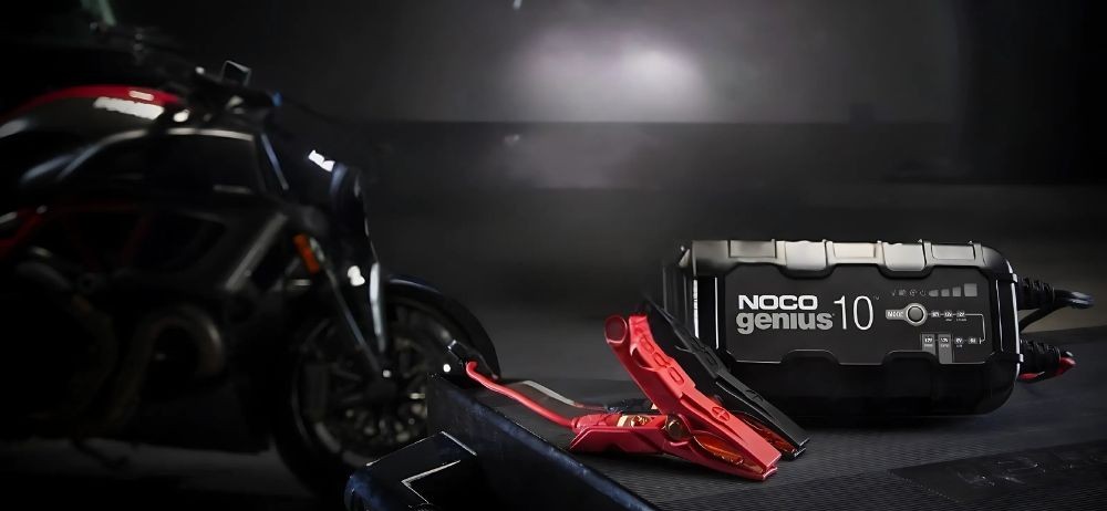 NOCO Genius 10 smart charger placed beside red battery clamps with a motorcycle in the background.