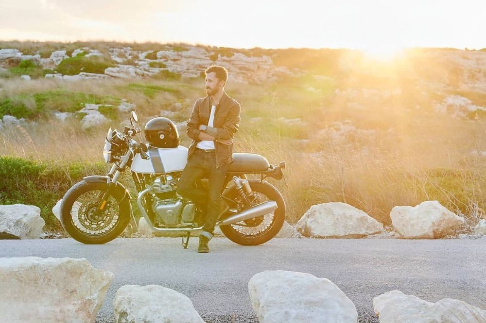 Motorcyclist in summer gear leaning on a parked bike at sunset on an open road with rocky terrain