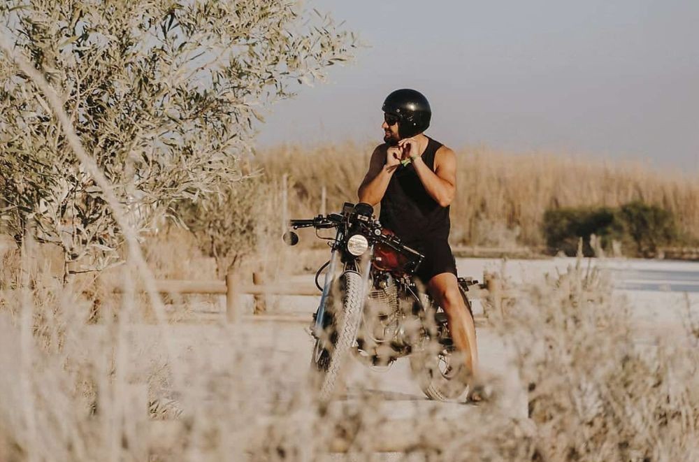 Man in minimal gear on a motorcycle adjusting helmet near a dry, rural trail on a sunny day
