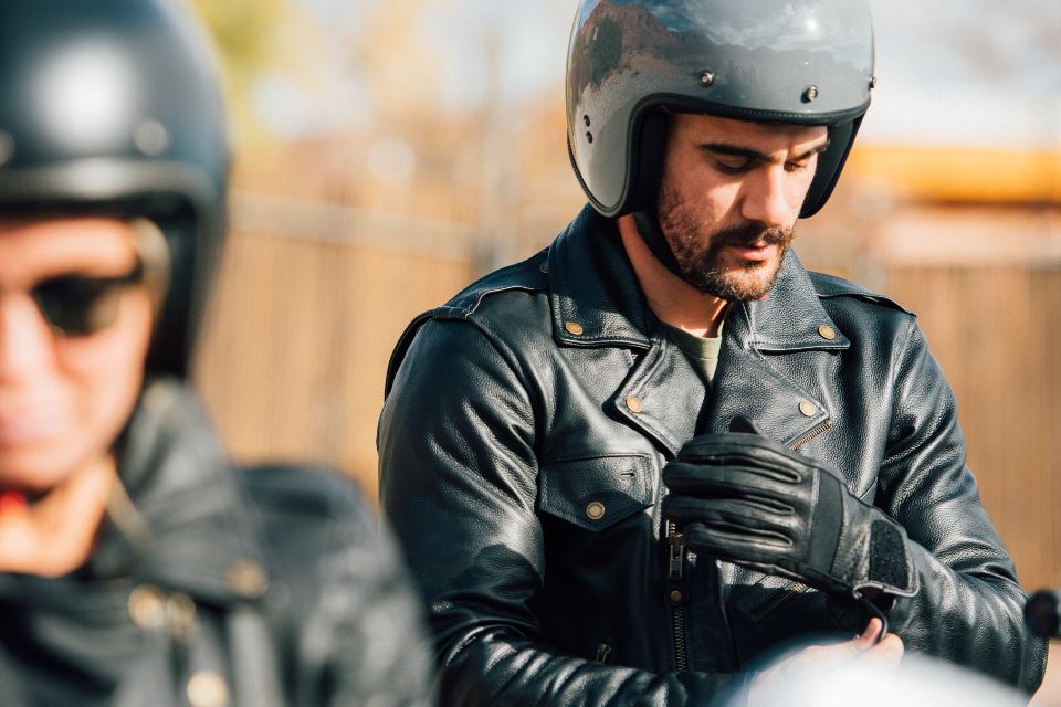 Motorcyclist wearing retro-style black leather gloves and jacket, adjusting his glove while seated outdoors in daylight.