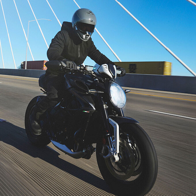 Motorcyclist wearing a matte modular helmet riding a naked bike across a bridge