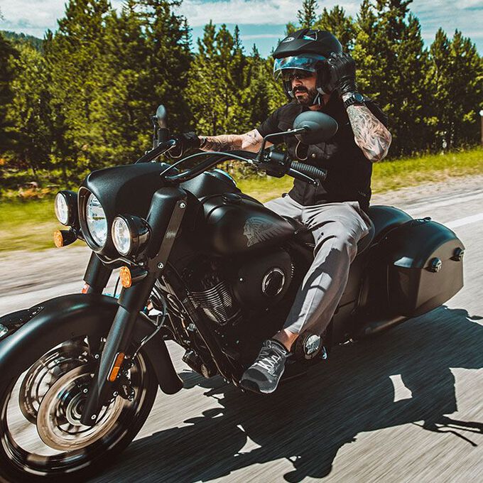 Rider on a cruiser motorcycle wearing sunglasses and an open-face helmet, riding through a forested highway