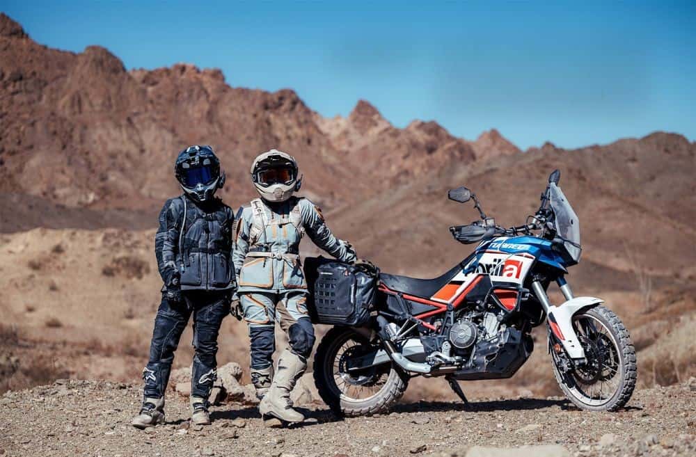 Two adventure riders in mesh jackets and pants pose next to a dual-sport bike in a dry, rocky landscape under clear blue skies.