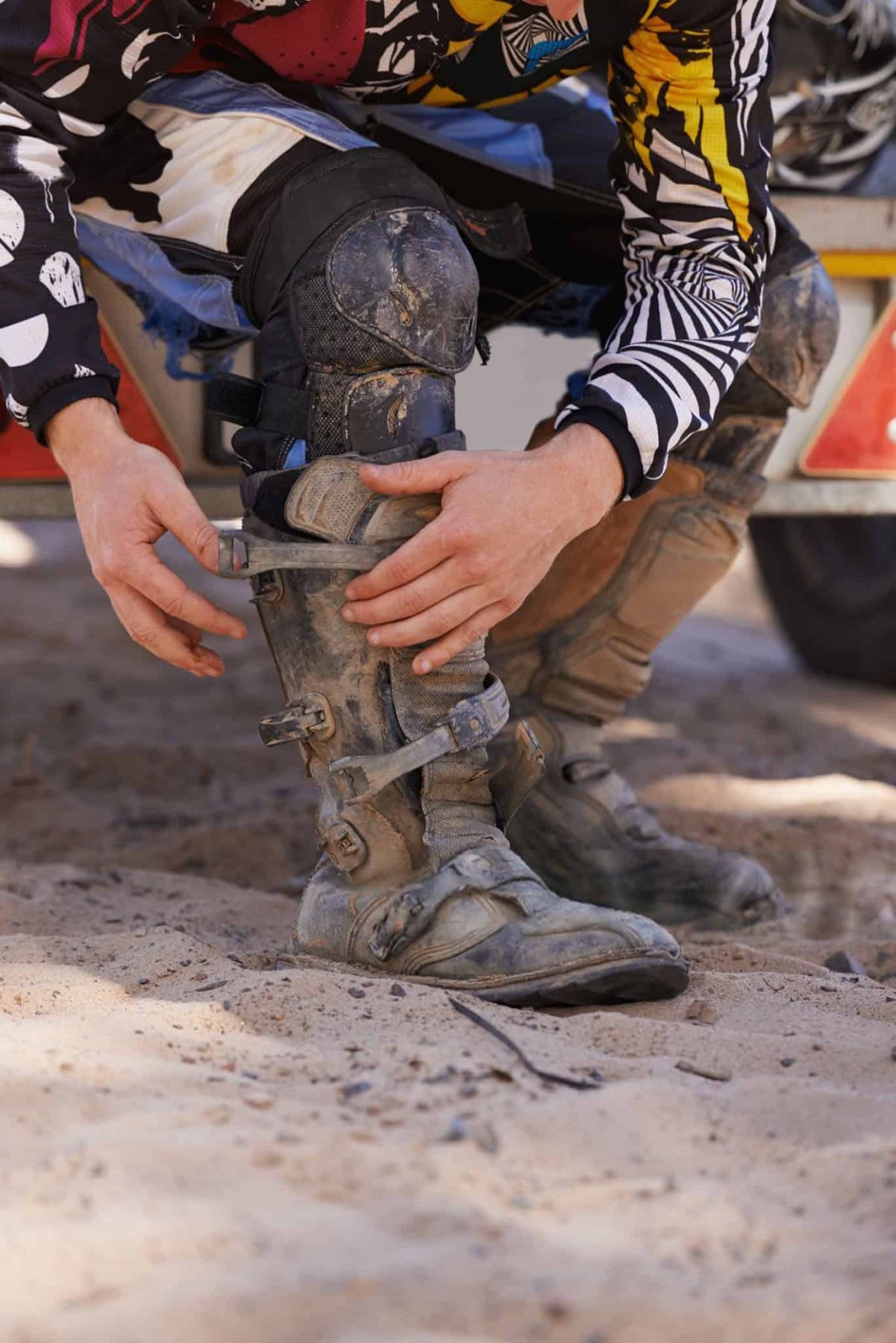 Close-up of a rider fastening motocross boots with worn, dusty buckles, highlighting real-world use and fit adjustment.