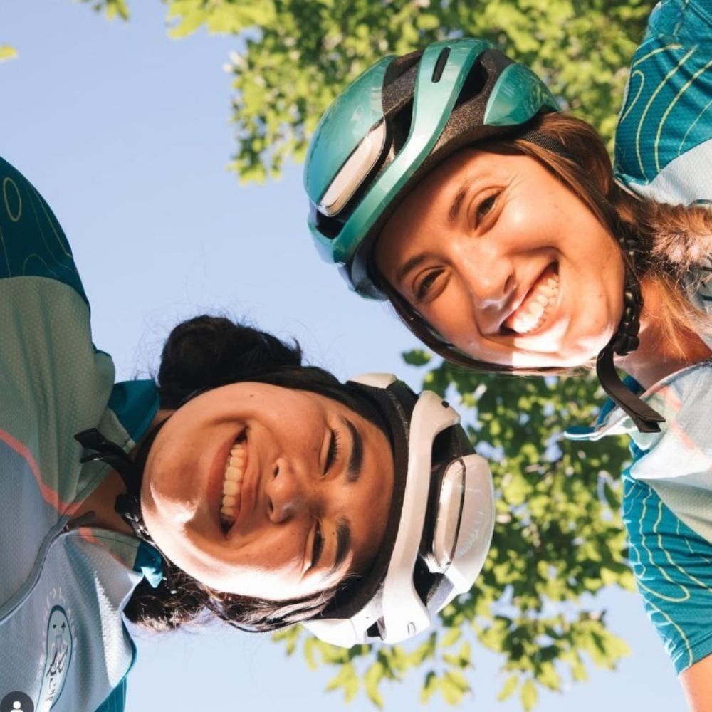 Two smiling women wearing colorful e-bike helmets, enjoying a sunny day outdoors under leafy trees