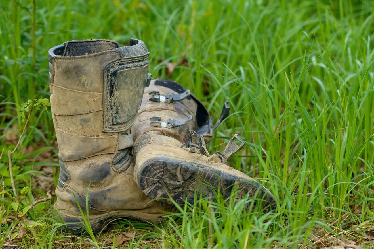 Muddy dual-sport boots resting in grass after a ride, showing the need for cleaning and care after off-road use.