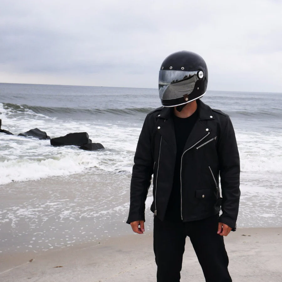 Rider wearing a First Manufacturing black leather motorcycle jacket and helmet, standing on a beach