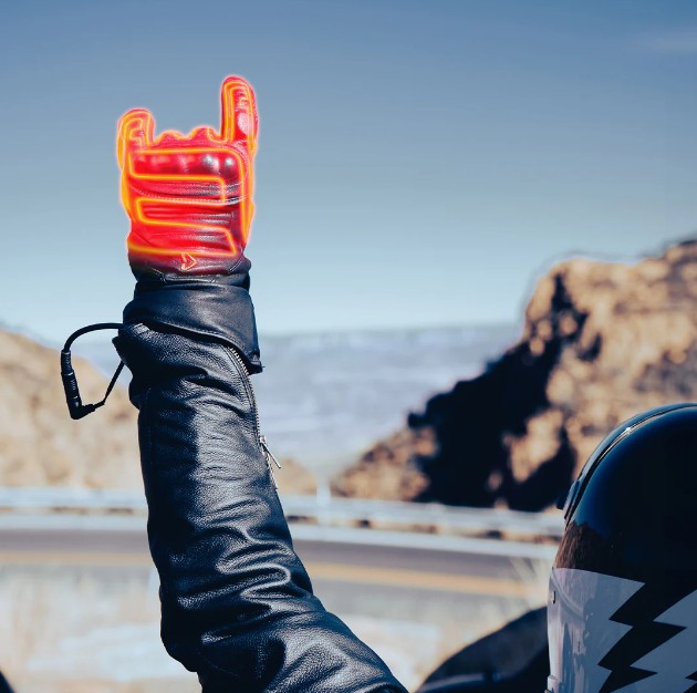 Motorcyclist wearing heated gloves with battery cable raised against a scenic mountain road background.