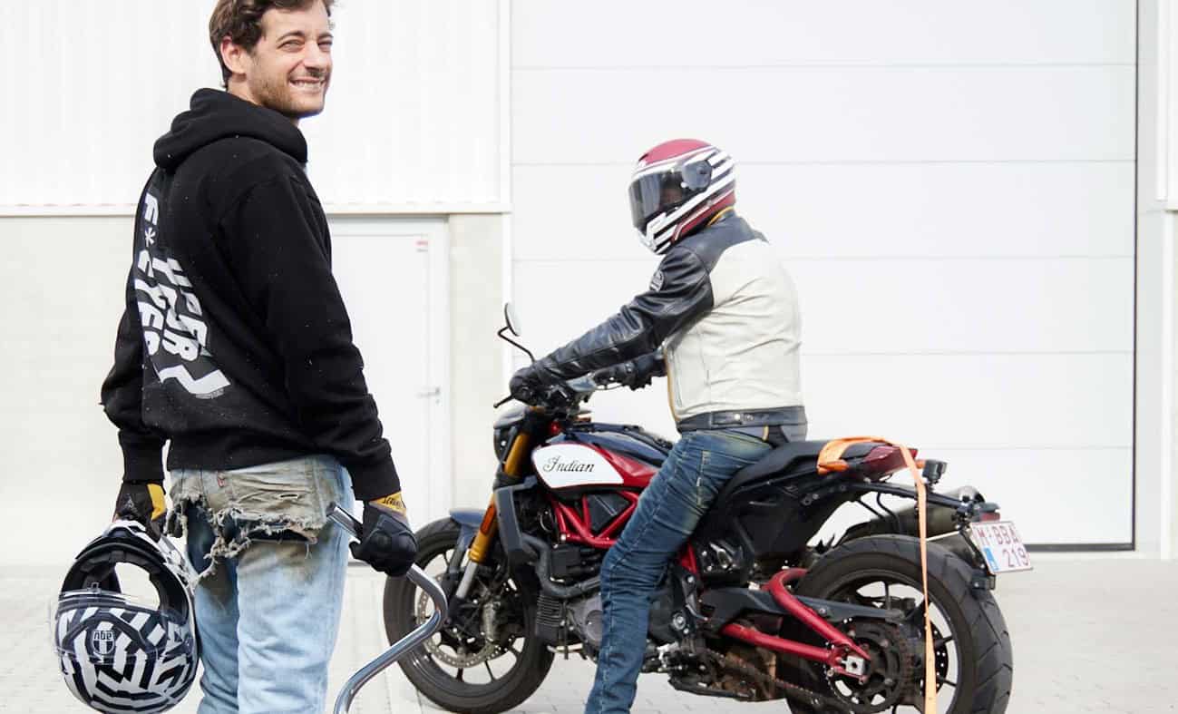Motorcyclist in casual jeans and hoodie gear beside another rider in protective clothing, both near a custom street bike